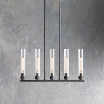 Five clear glass cylinders on a black linear chandelier against a gray wall.