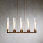 Five clear glass cylinders on a brass-tone linear chandelier against a gray wall.