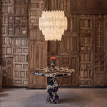 Illuminated tiered selenite chandelier above a pedestal table against a wood-paneled wall.