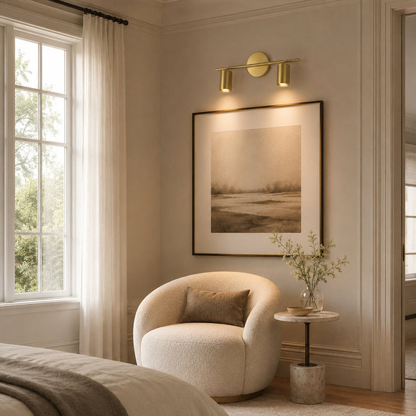 Twin-head brass picture light illuminating framed artwork in a warm bedroom sitting area