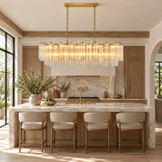 Layered linear glass chandelier above a pale kitchen island with four upholstered stools.