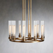 Brass-tone ring chandelier with upright clear glass cylinders against a gray wall.